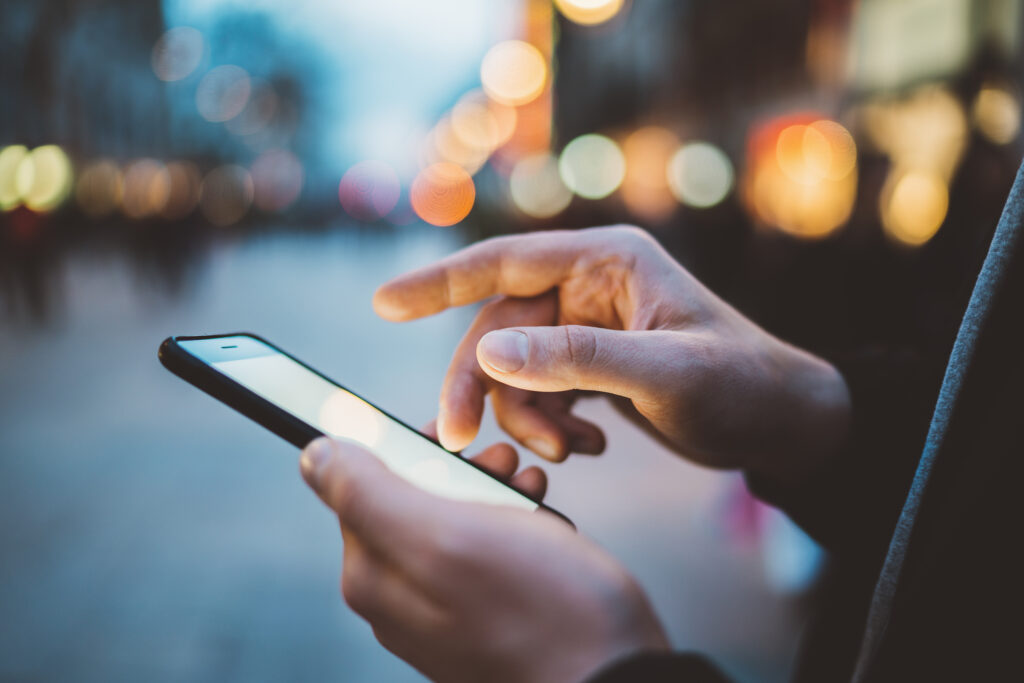 Close-up image of male hands using smartphone at night on city shopping street, searching or social networks concept, hipster man typing an sms message to his friends